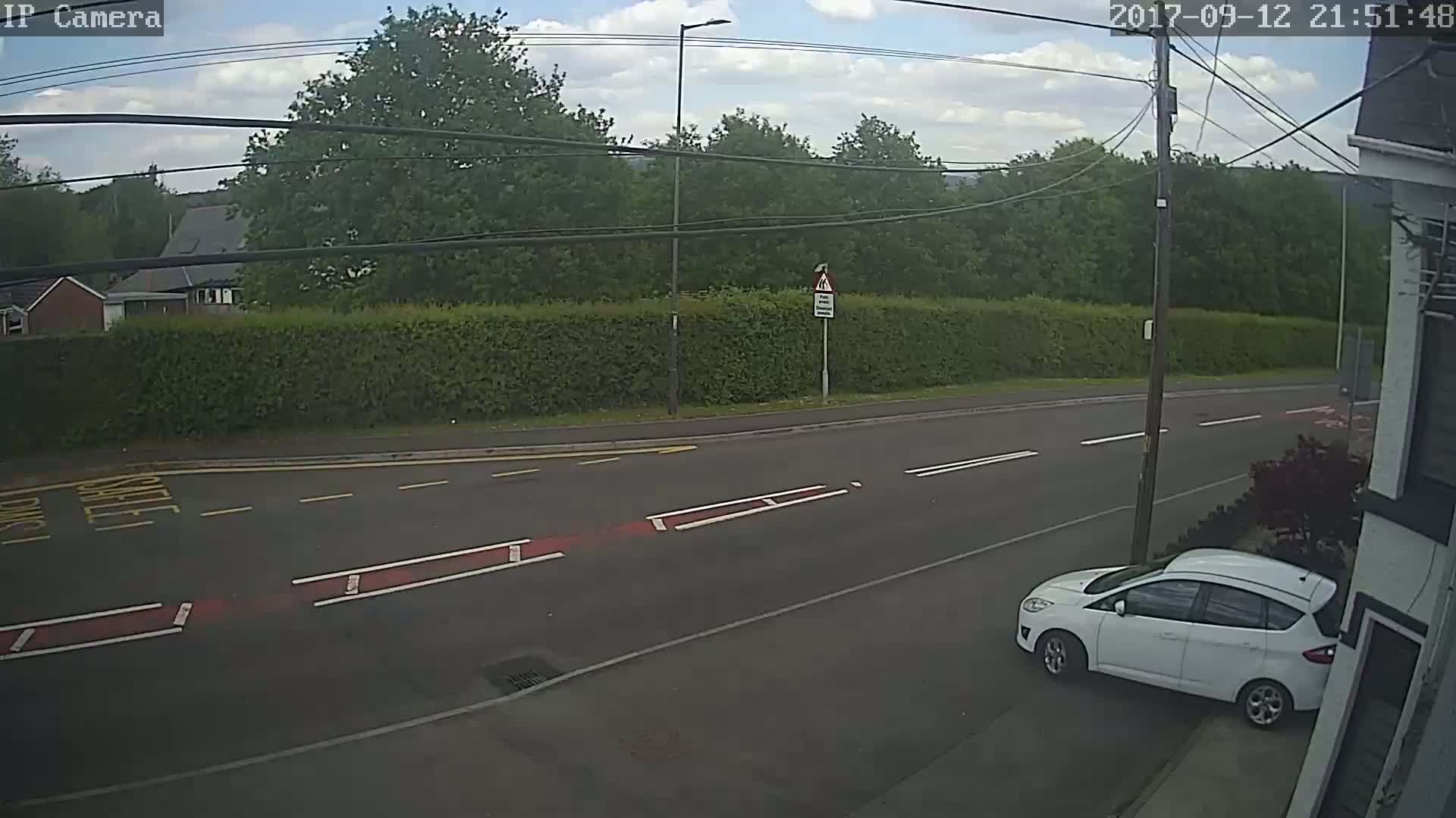 A partly cloudy day shows a white car parked on the side of a road, beside a tall hedge and some trees, under a mostly clear sky.