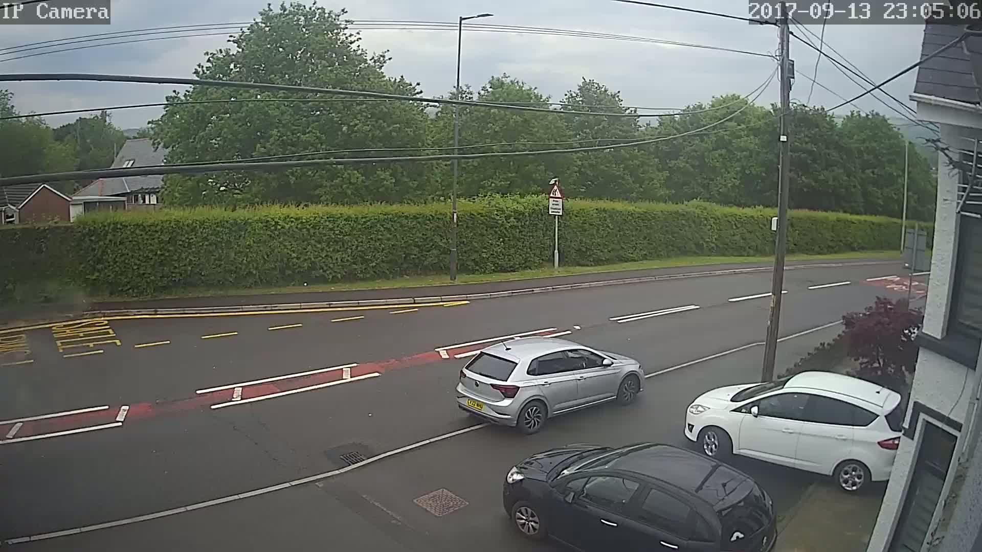 A gray car and a white car are parked on the side of a wet road lined with green hedges and trees under an overcast sky.