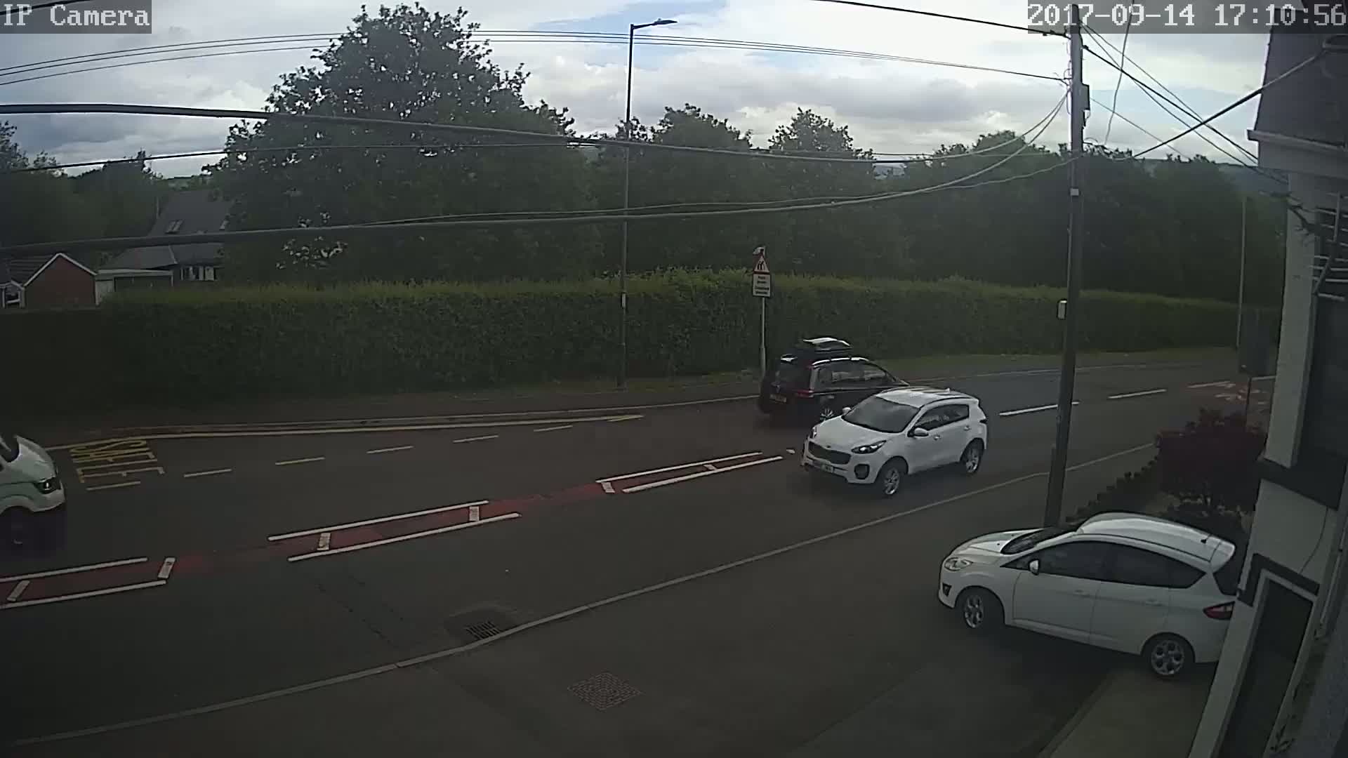 A partly cloudy day shows three cars on a road lined with hedges and trees.