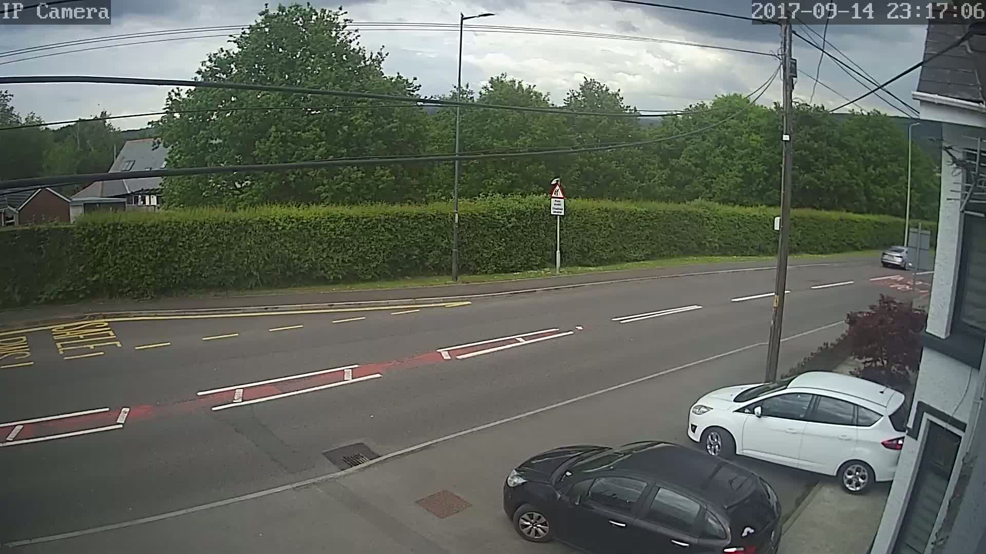 A mostly cloudy day shows a two-lane road with two parked cars, one white and one black, near a tall hedge and some buildings in the distance.