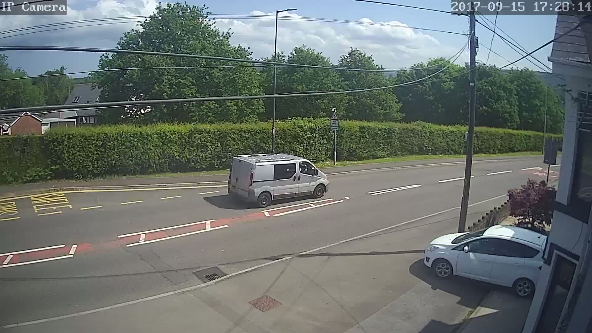 A light-grey van and a white car are visible on a road under a partly sunny sky.
