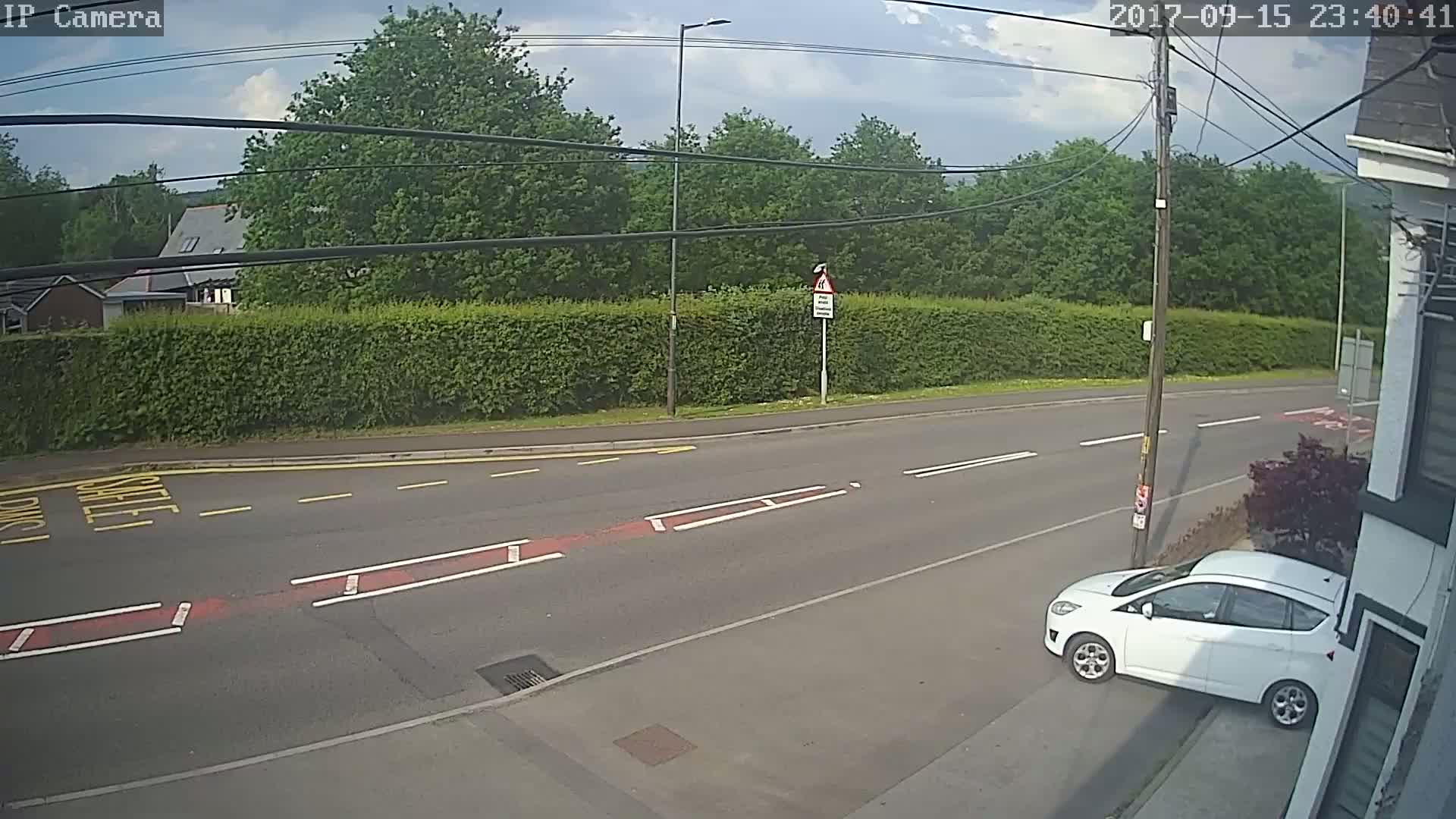 A white car is parked on the side of a road, which is lined with a tall green hedge and trees under a partly cloudy sky.