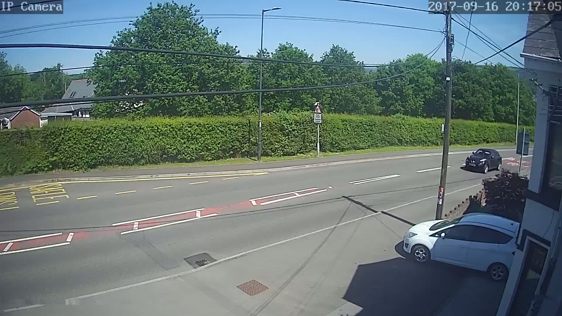 A sunny day shows a road intersection with two cars, one dark-colored and one white, and a green hedge behind it.