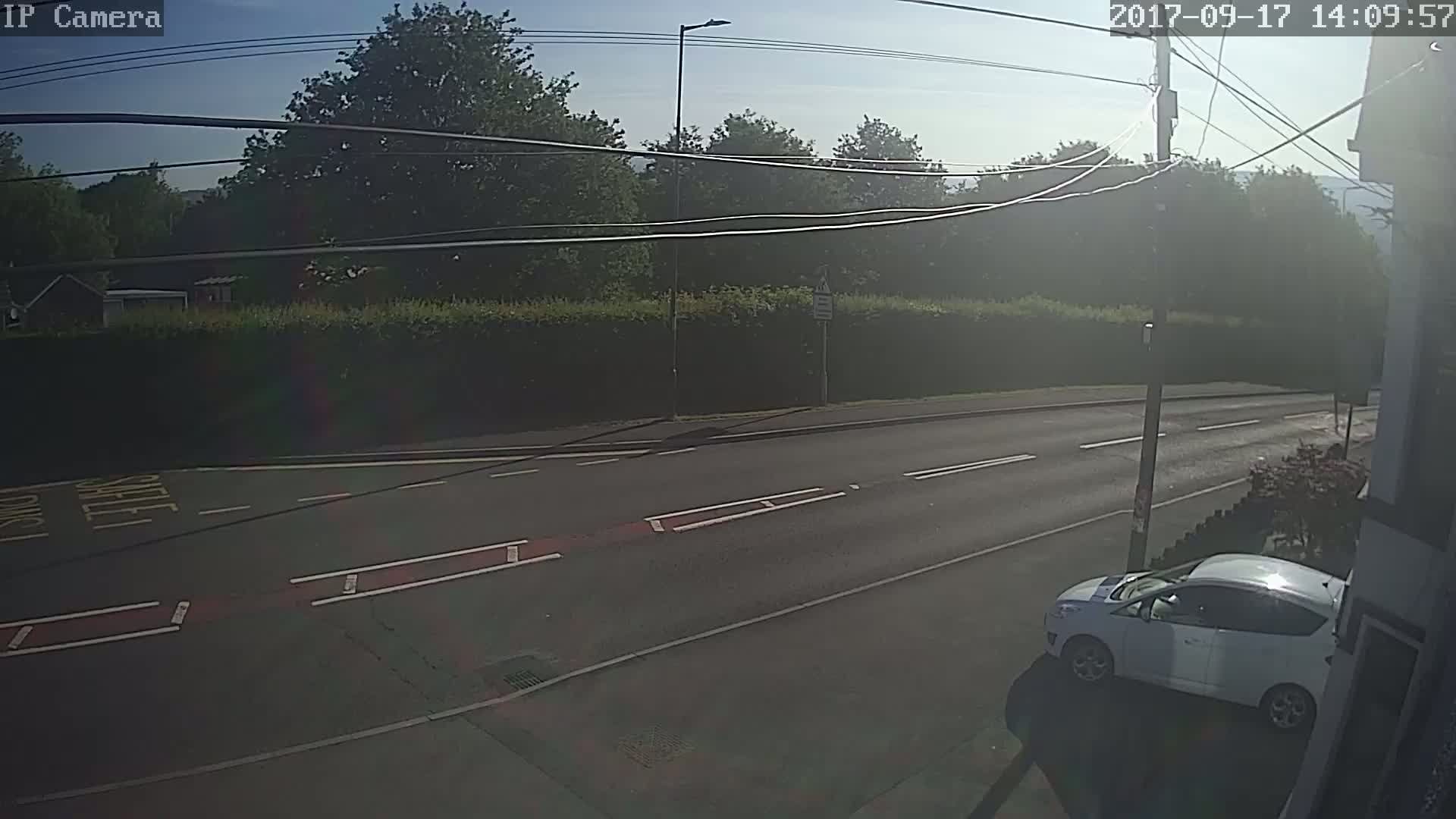 A sunny day shows a road with a white car parked on the roadside, next to a hedgerow and trees beyond.
