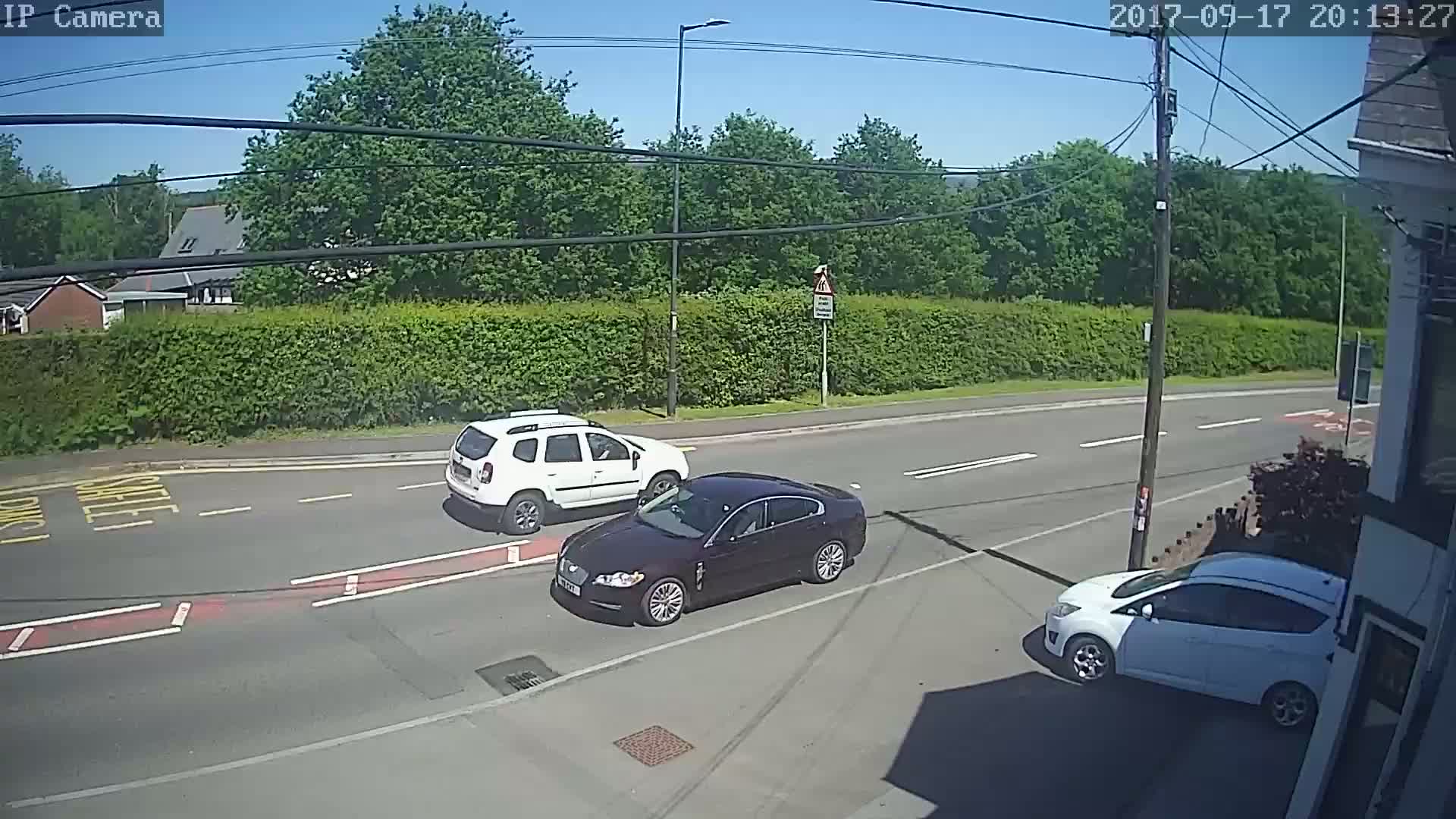 On a sunny day, three cars are parked or slowly moving on a road bordered by a green hedge, with trees and buildings in the background.