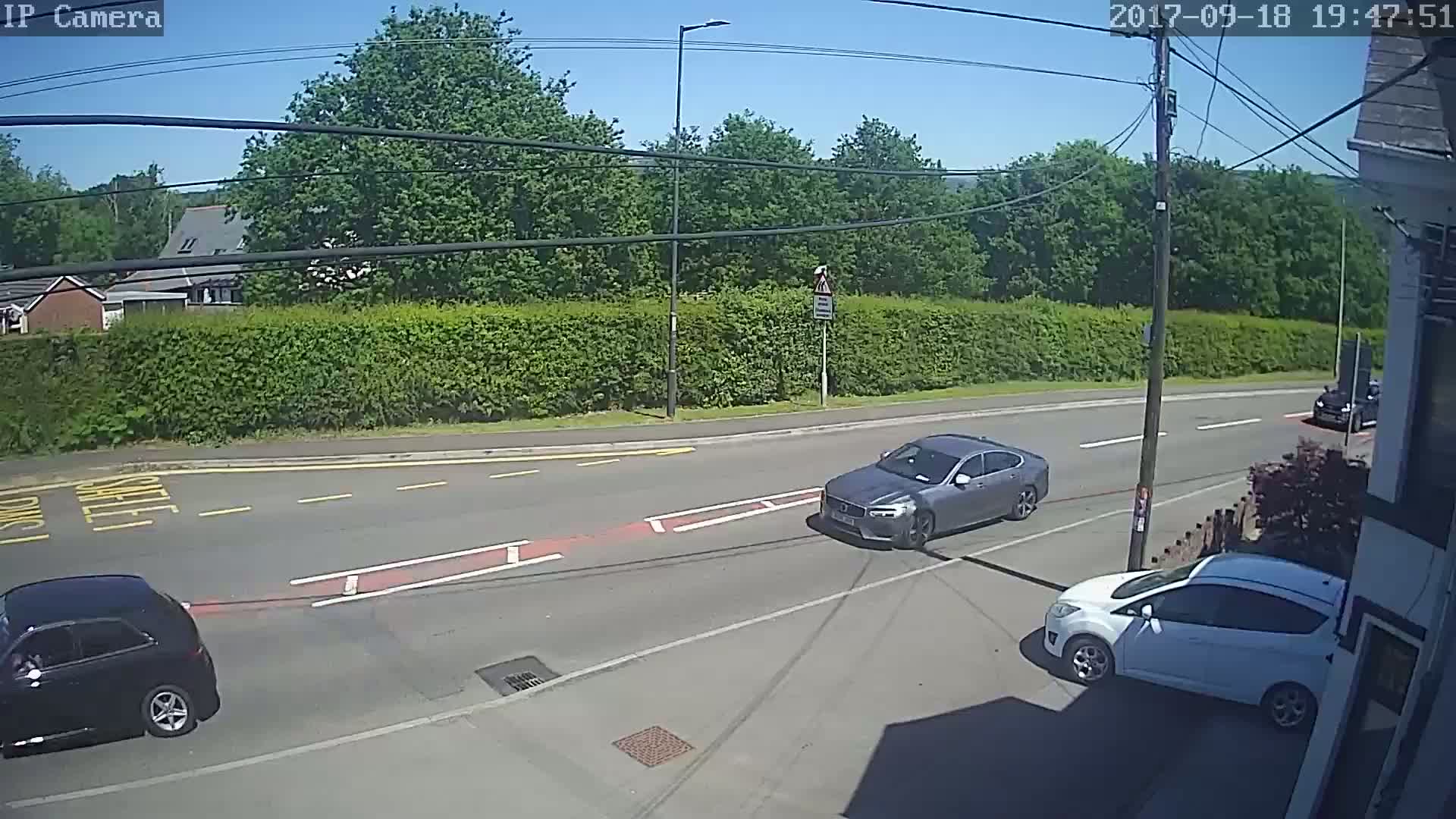 A sunny day shows three cars on a road next to a tall hedge, with houses and trees visible in the background.
