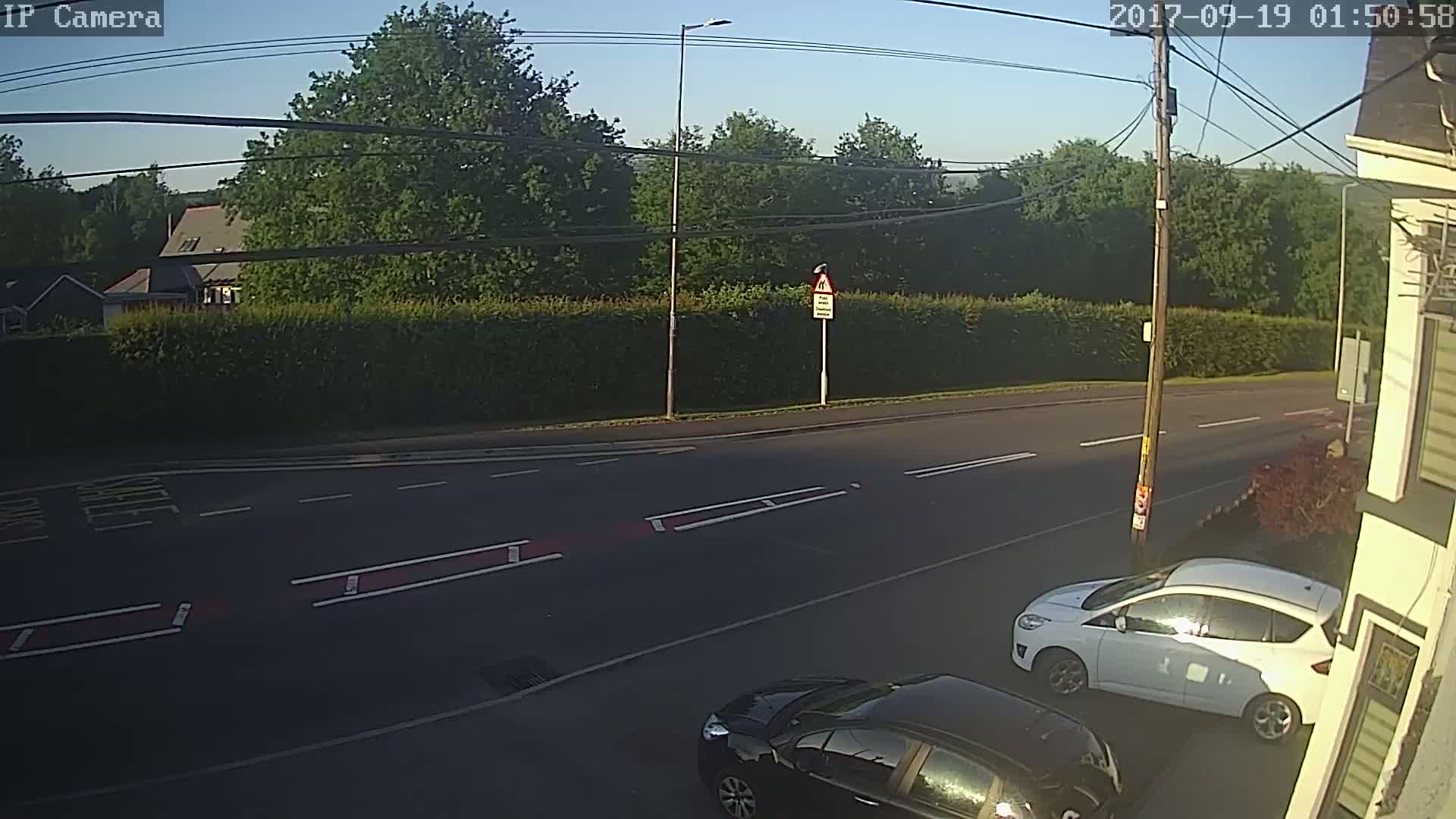 A sunny morning view of a road with two cars parked, one white and one black, near a hedge and trees.