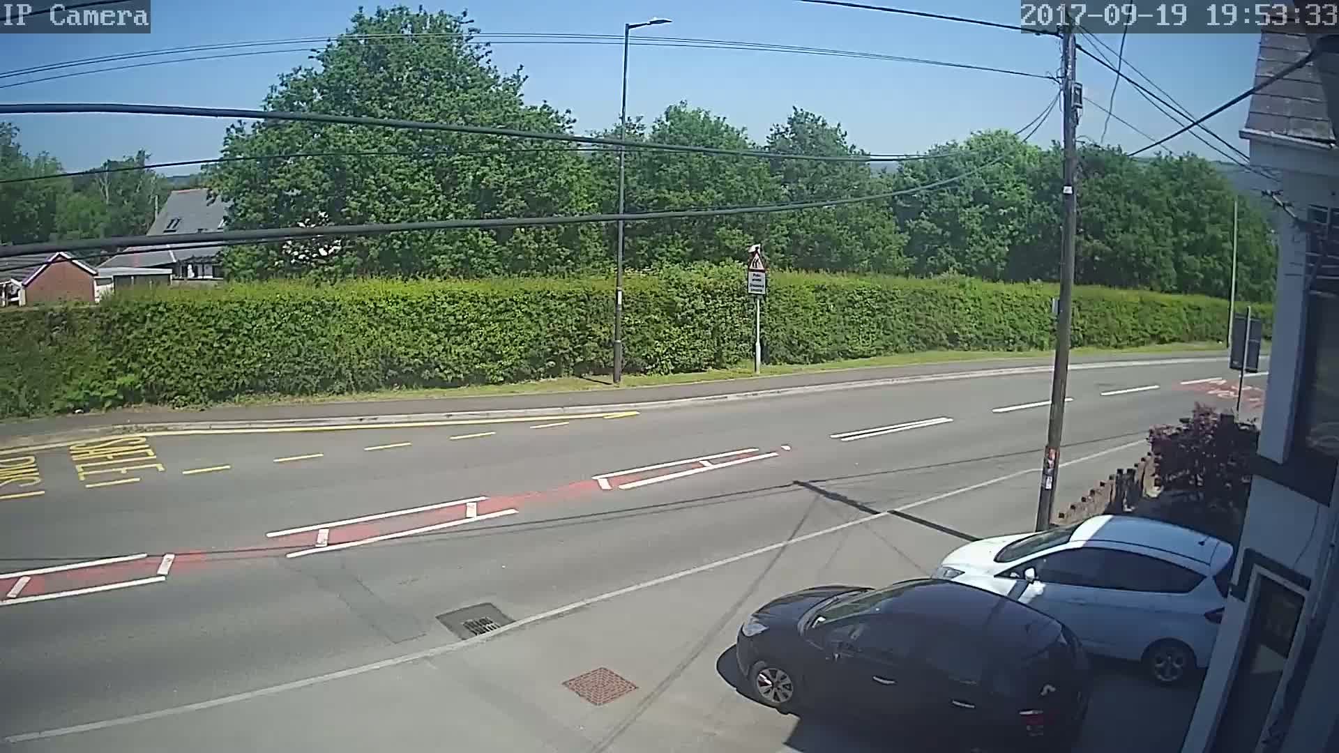 A sunny view of a road intersection with two cars parked, bordered by a tall hedge and trees under a clear sky.