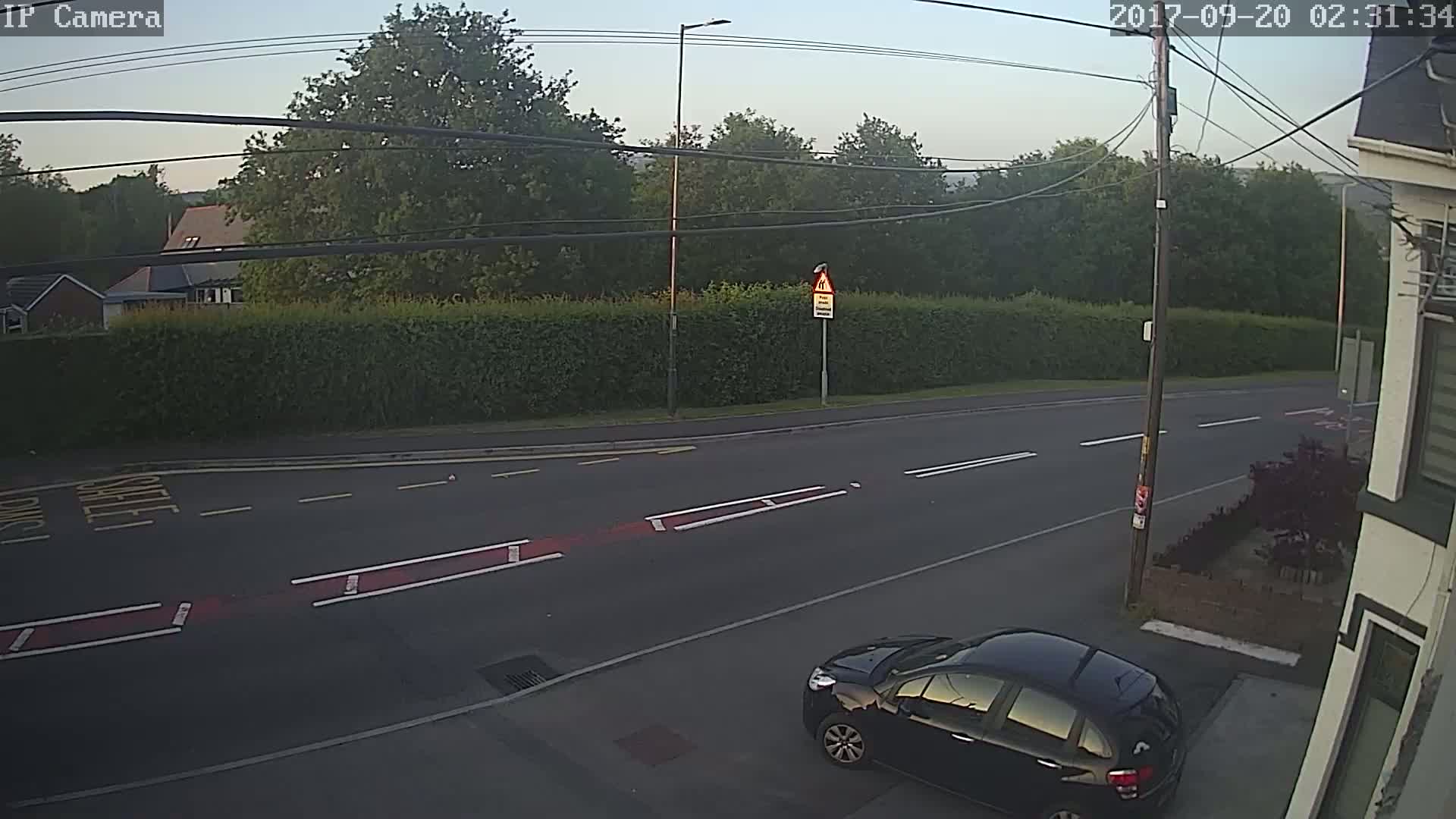 A black car is parked on the side of a road, next to a hedgerow and trees under a clear, light sky.