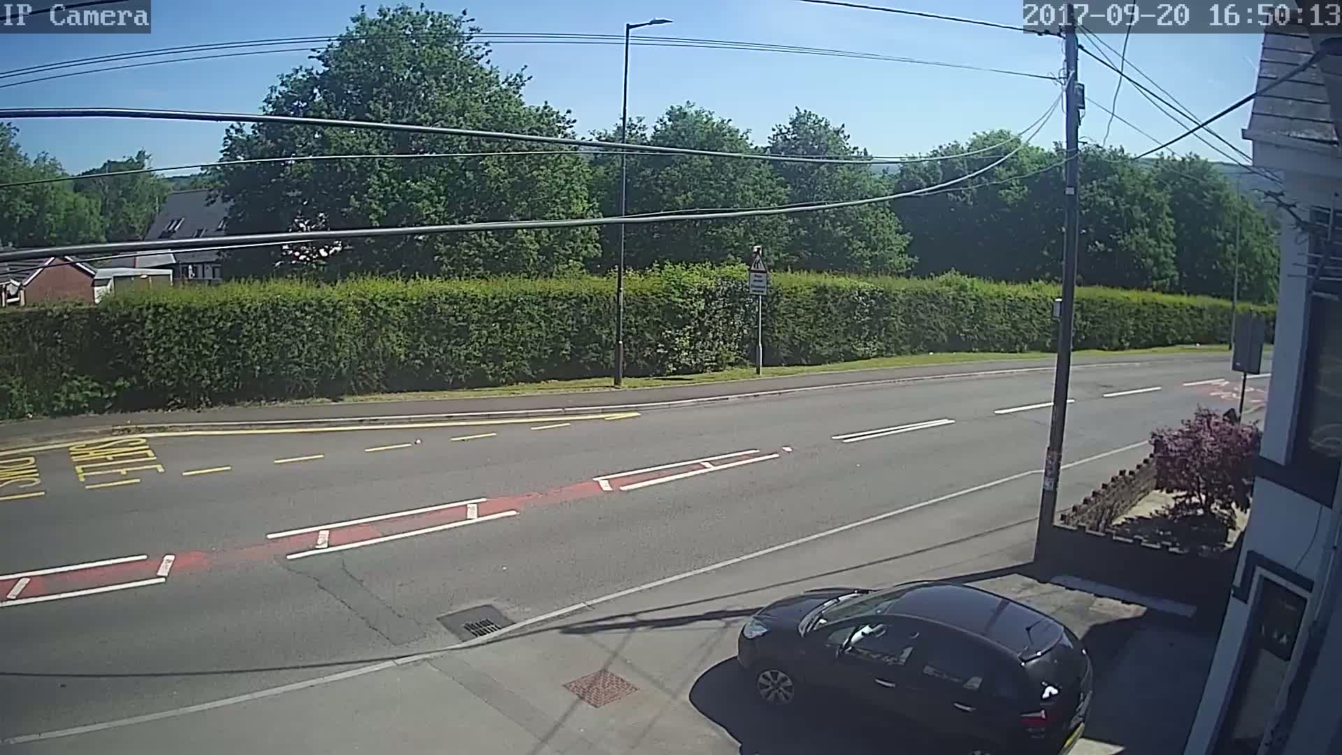 A sunny day shows a dark-colored car parked near a road with a hedge and trees in the background.