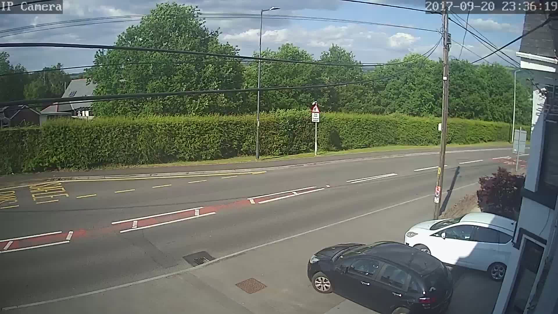 A partly sunny day shows a road with two cars parked on the side, a hedge, and trees in the background.