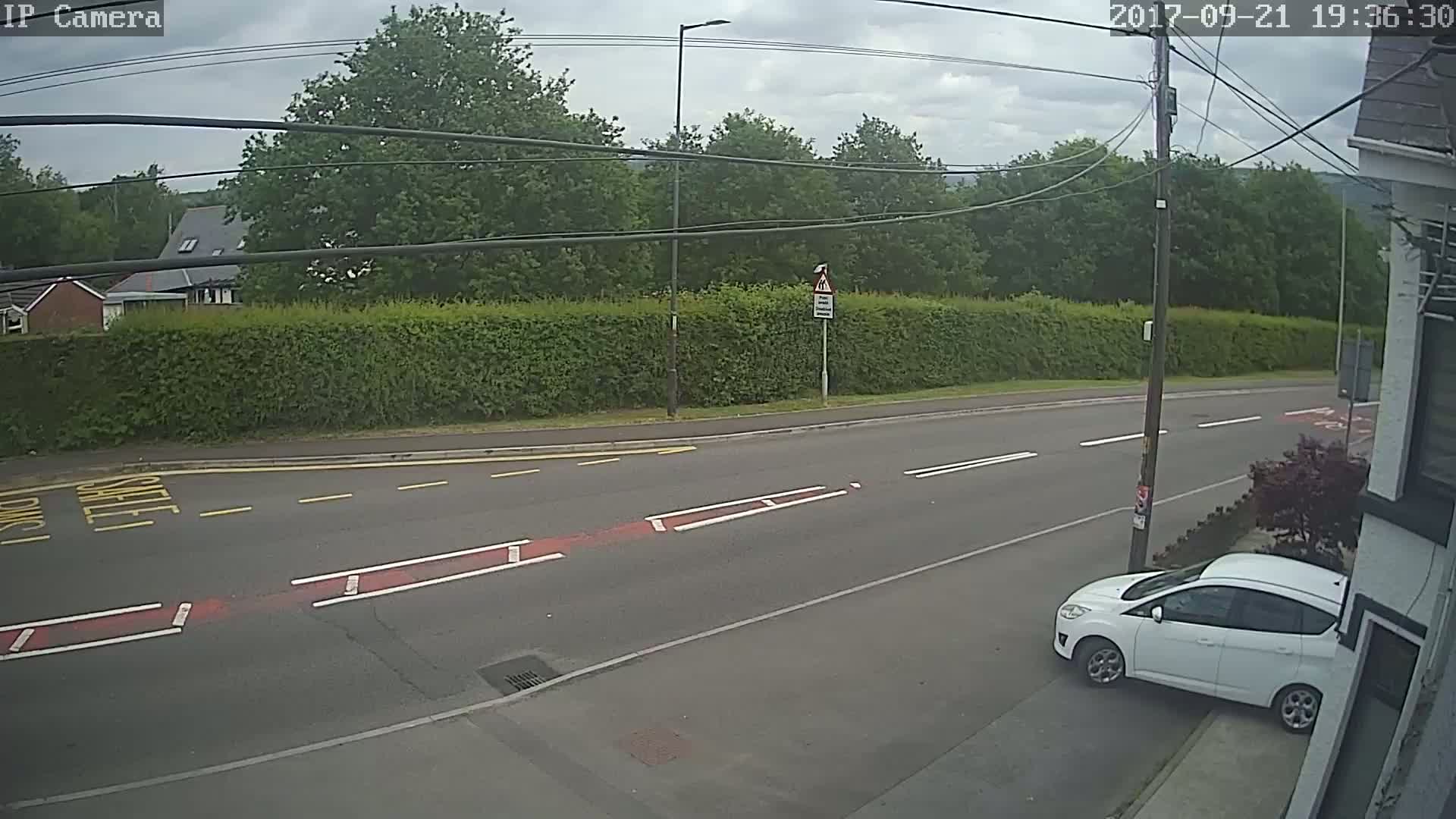 A white car is parked on the side of a road, which is bordered by a tall hedge and trees under a partly cloudy sky.