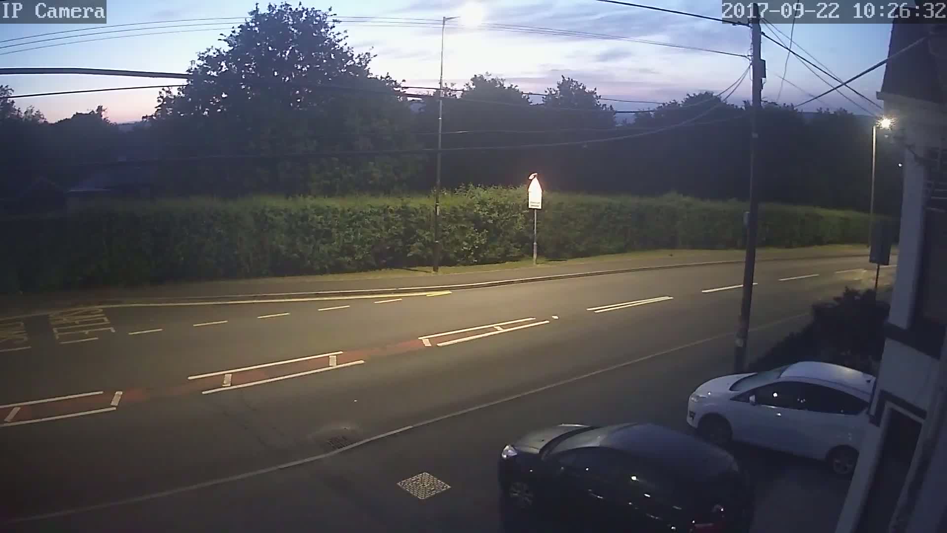 A twilight view of a road with two cars parked, one white and one dark-colored, alongside a hedgerow and trees under a partly cloudy sky.