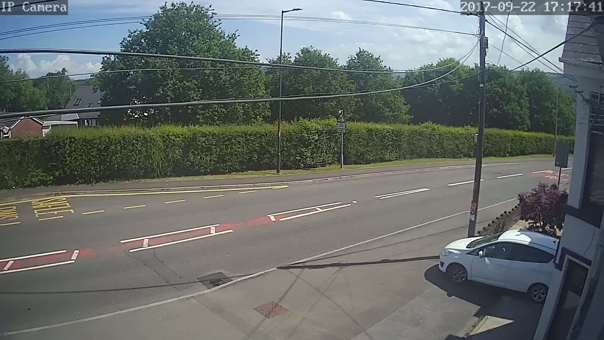 A sunny day shows a white car parked on the side of a road, across from a tall hedge and some trees.