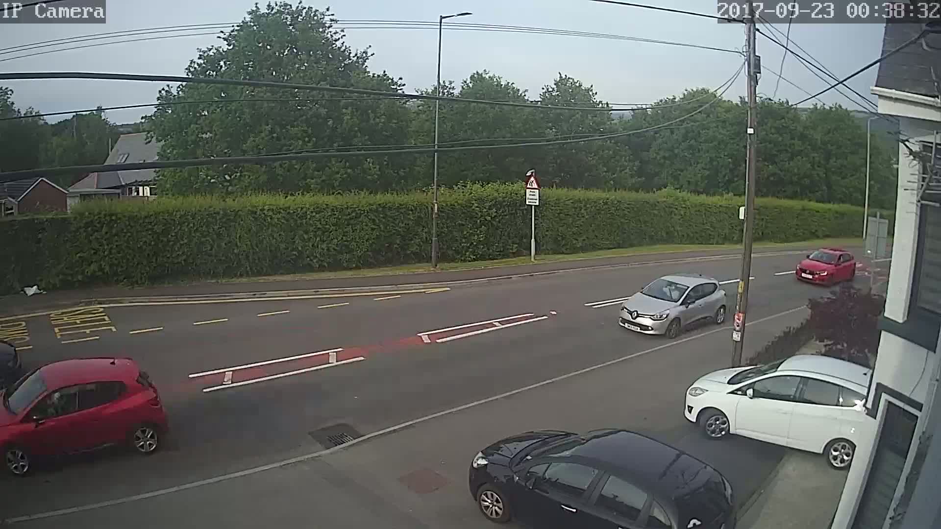 Several cars are driving on a two-lane road lined with green hedges under an overcast sky.