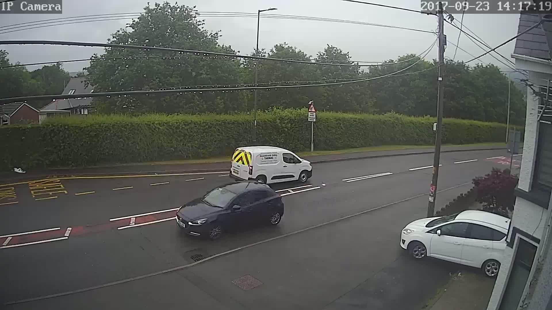 A rainy day shows a dark-colored car and a white van driving on a street, with another white car parked on the side.