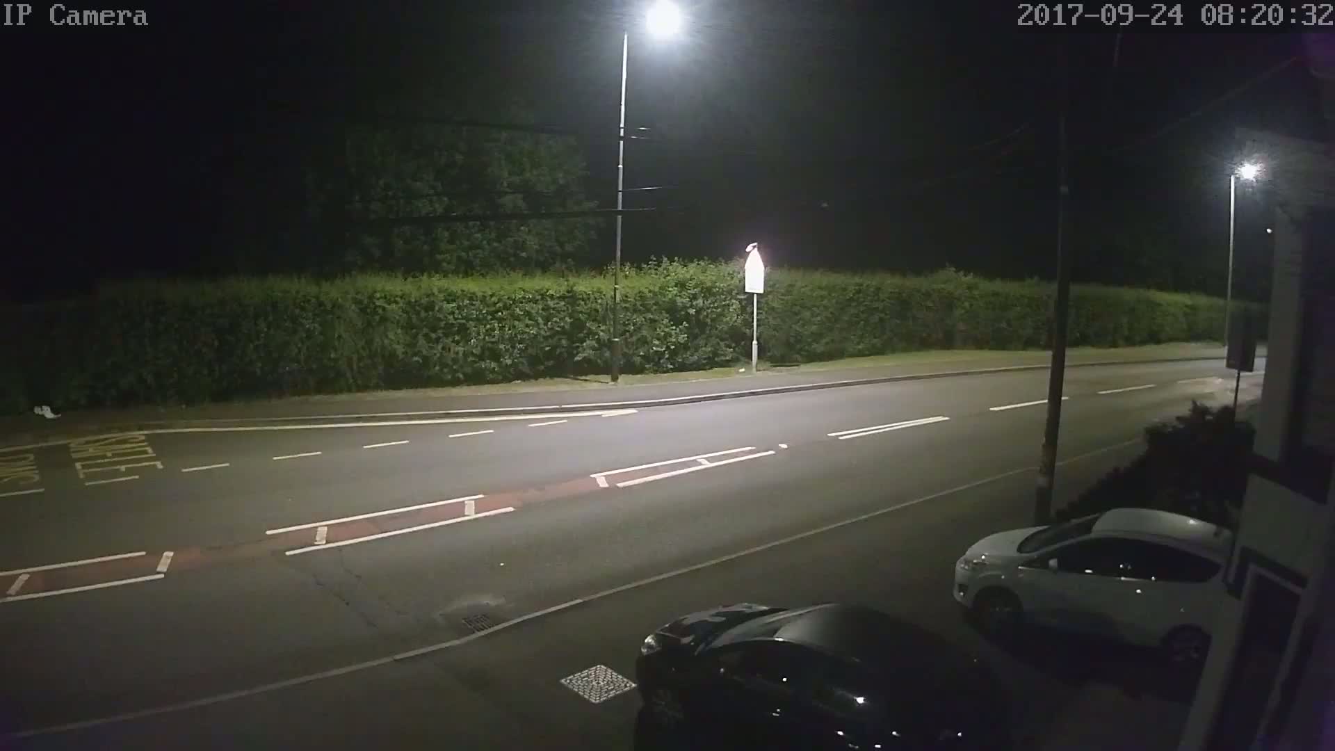 A nighttime street scene shows a road with two cars parked, one white and one dark-colored, next to a hedge under streetlights.