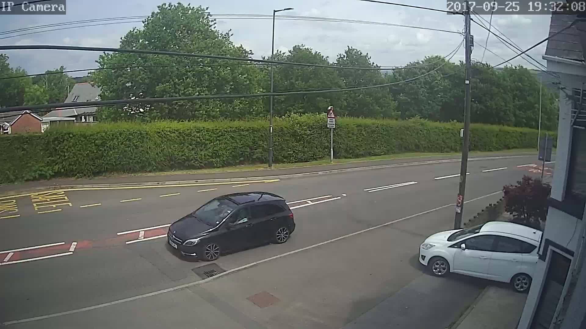 A black car drives past a white car parked on the side of a road under a partly cloudy sky.