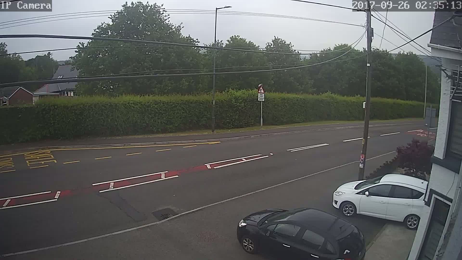 A mostly overcast day shows a residential street with two parked cars, one white and one black, next to a hedge, across from a road with painted traffic markings.
