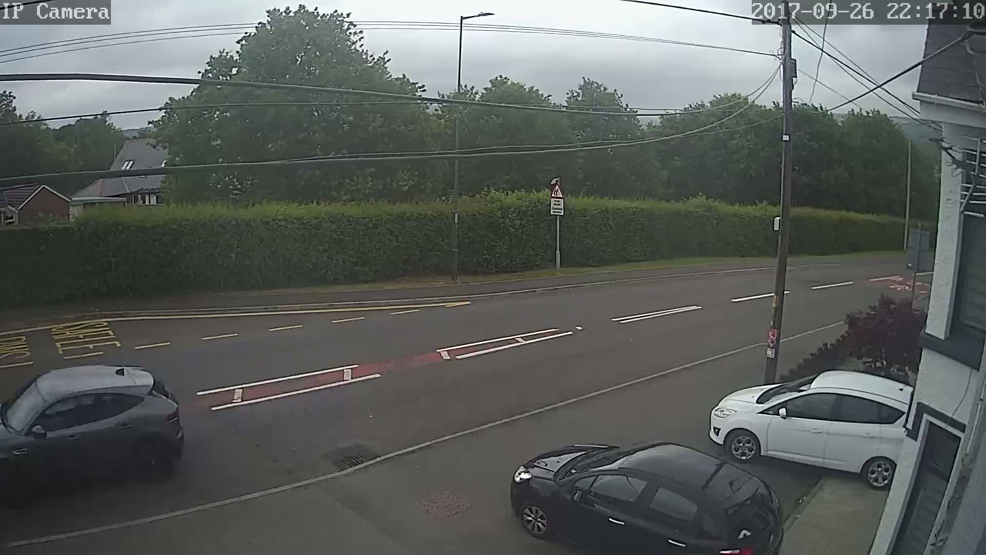 A gray car and a black car are parked on a residential street under an overcast sky, near a white car parked on the side of the road.