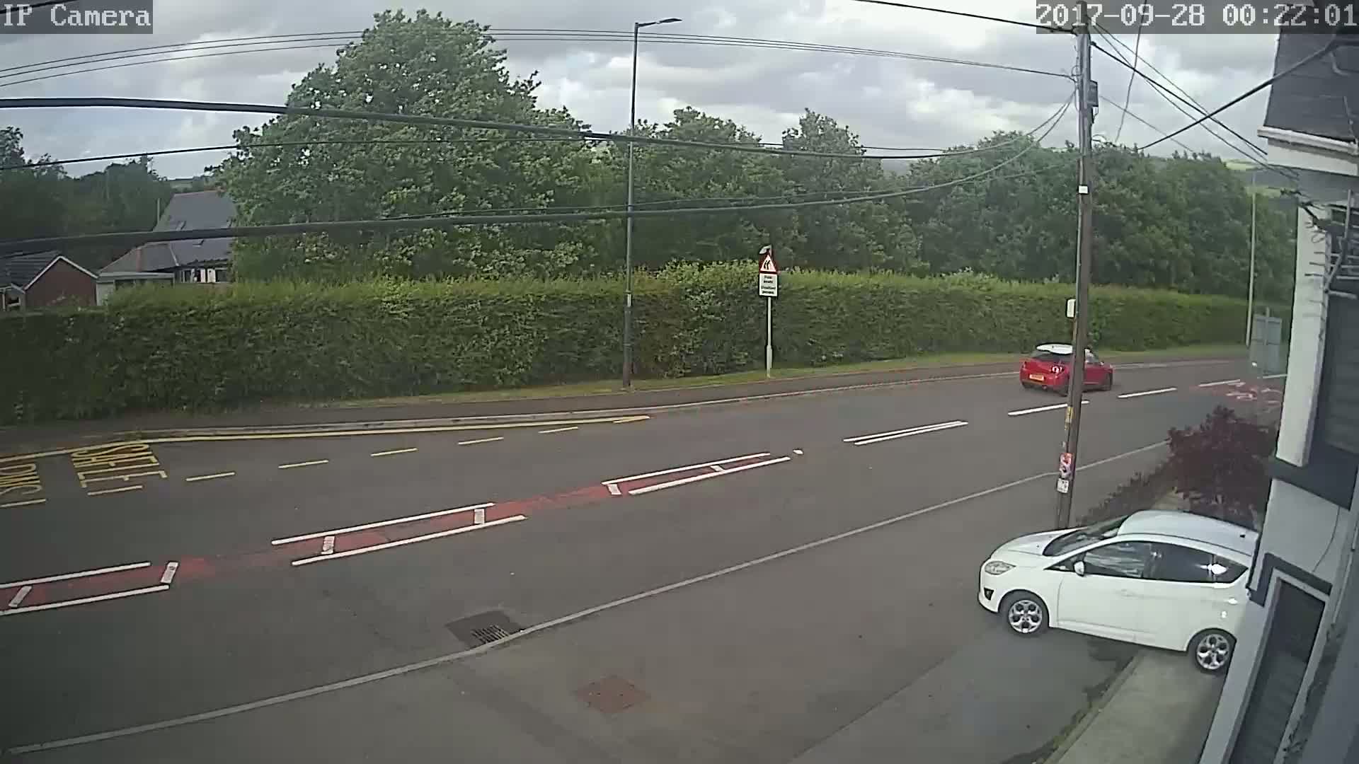 A cloudy day shows a road with a red car and a white car, one driving and one parked, with lush green hedges and trees in the background.