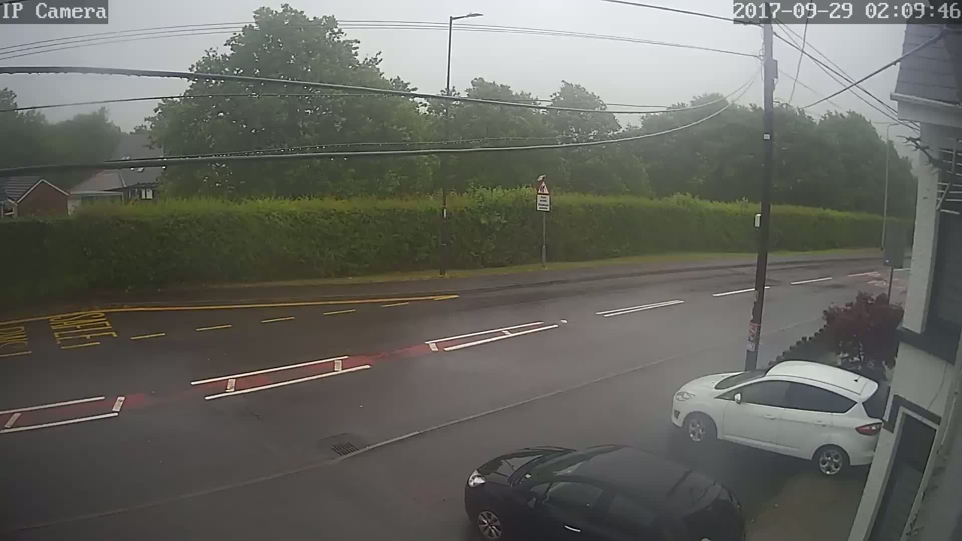 A rainy scene shows a wet road with two cars parked, one white and one dark-colored, next to a hedge and trees in the background.