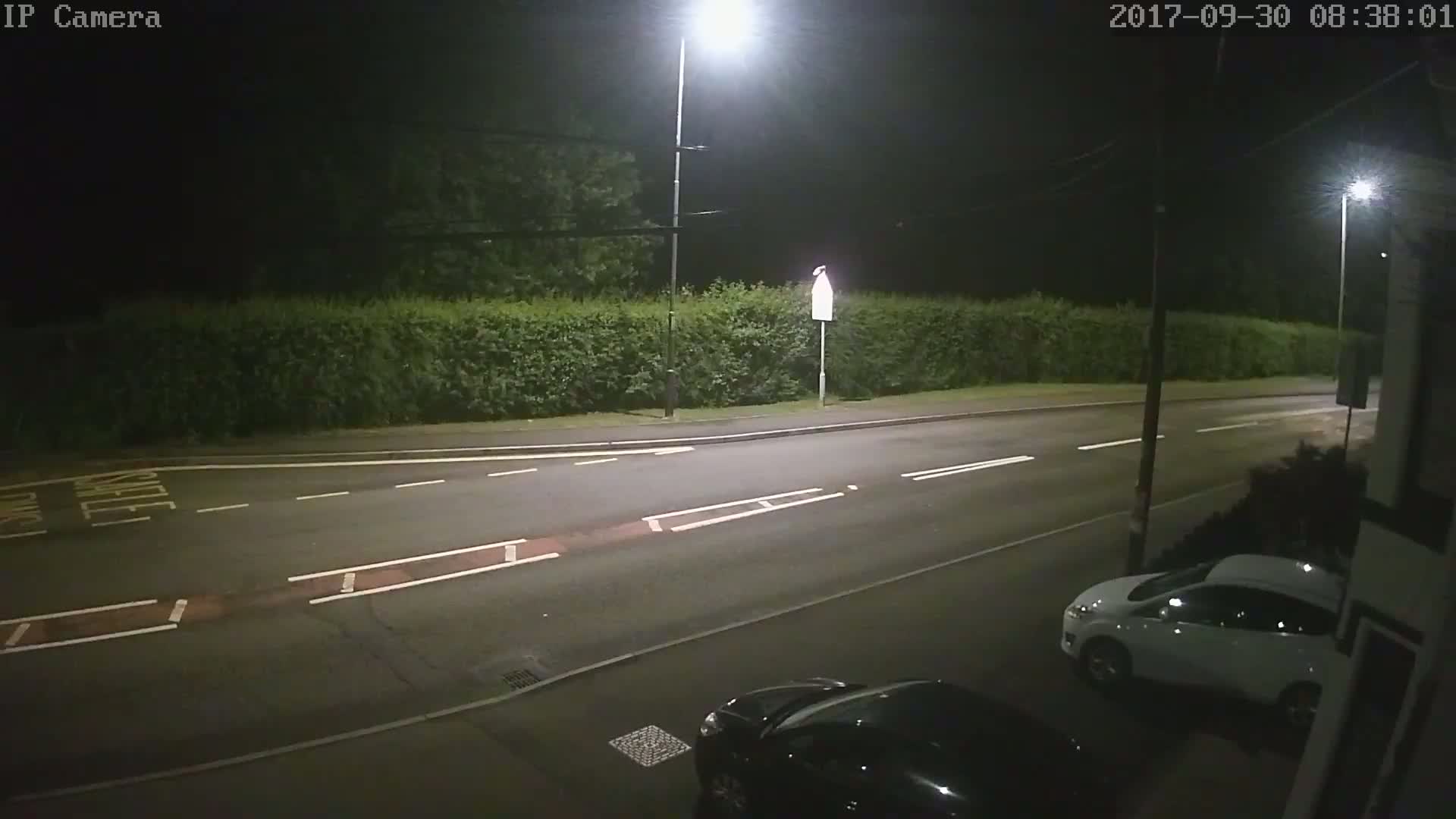 A nighttime view of a mostly empty road with a white car parked on the right and a dark car partially visible at the bottom, illuminated by streetlights, bordered by a hedge.