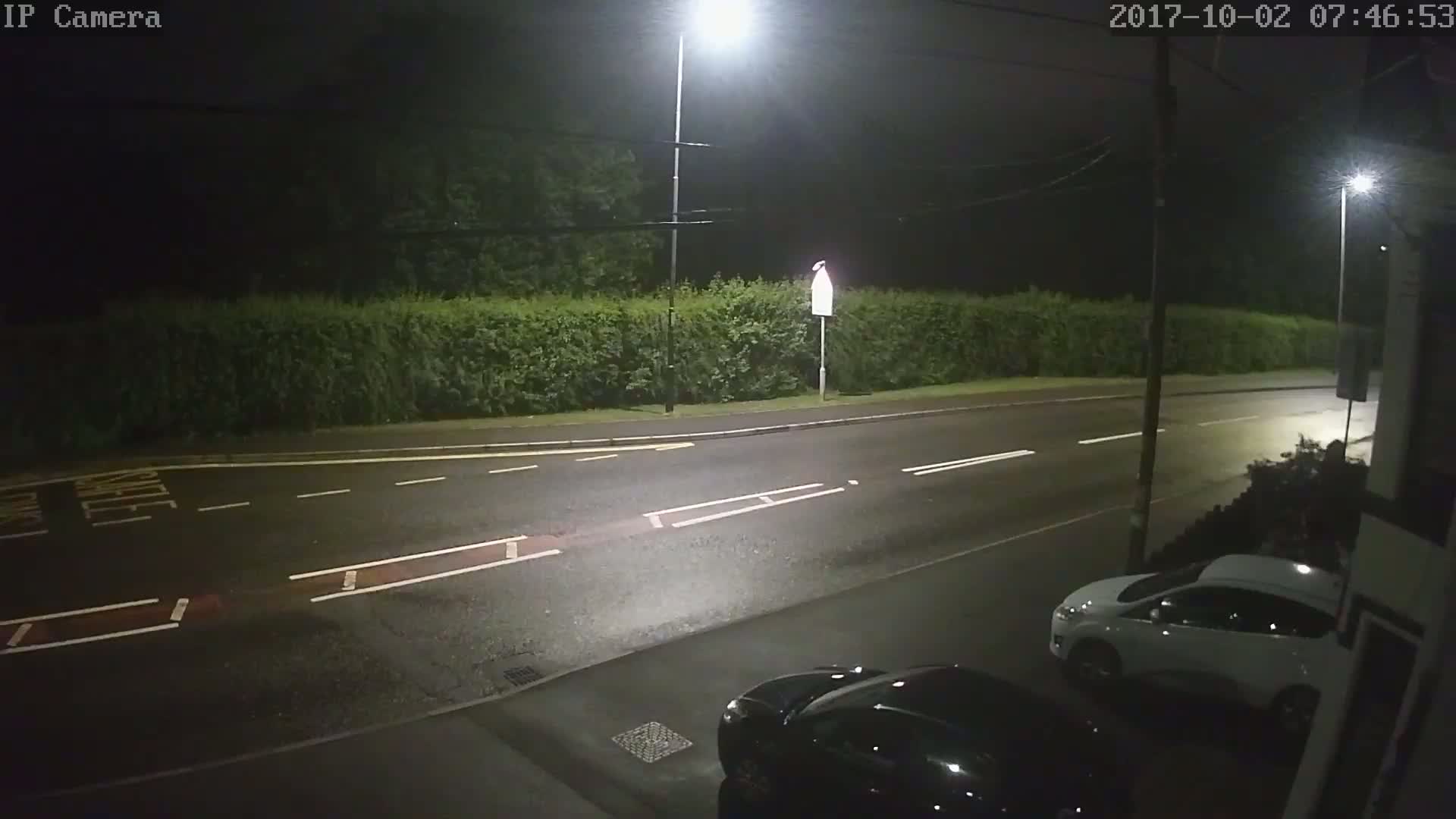 A wet, dark street at night is lit by streetlights, showing a white car parked and a dark car partially visible in the foreground.