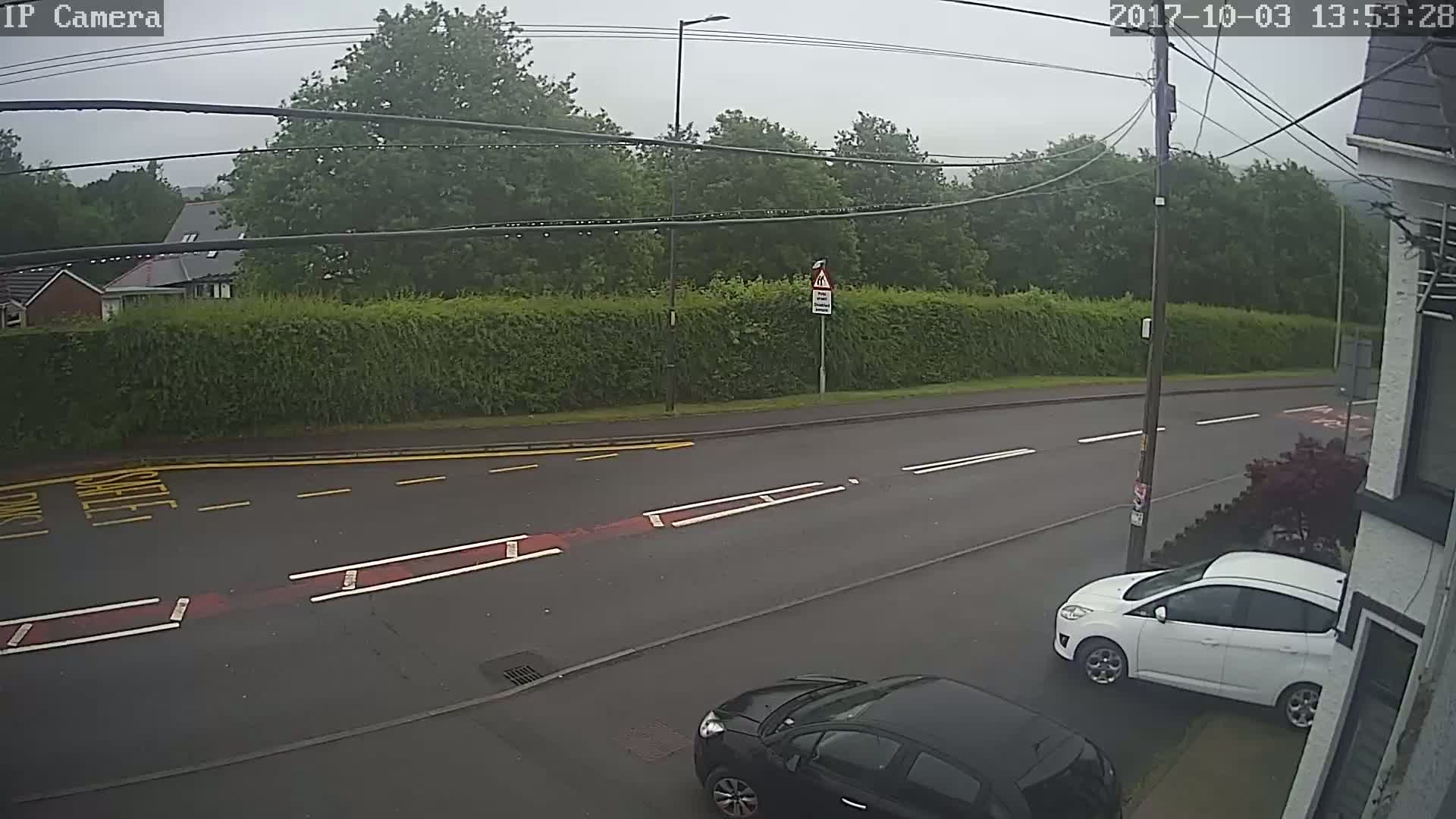 A rainy day shows a residential street with two cars parked, one white and one black, and a tall hedge separating the road from a row of houses.