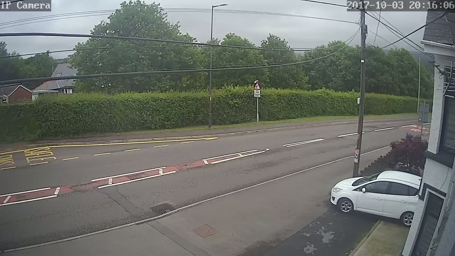 A white car is parked on the side of a road, which is bordered by a hedge and trees under an overcast sky.