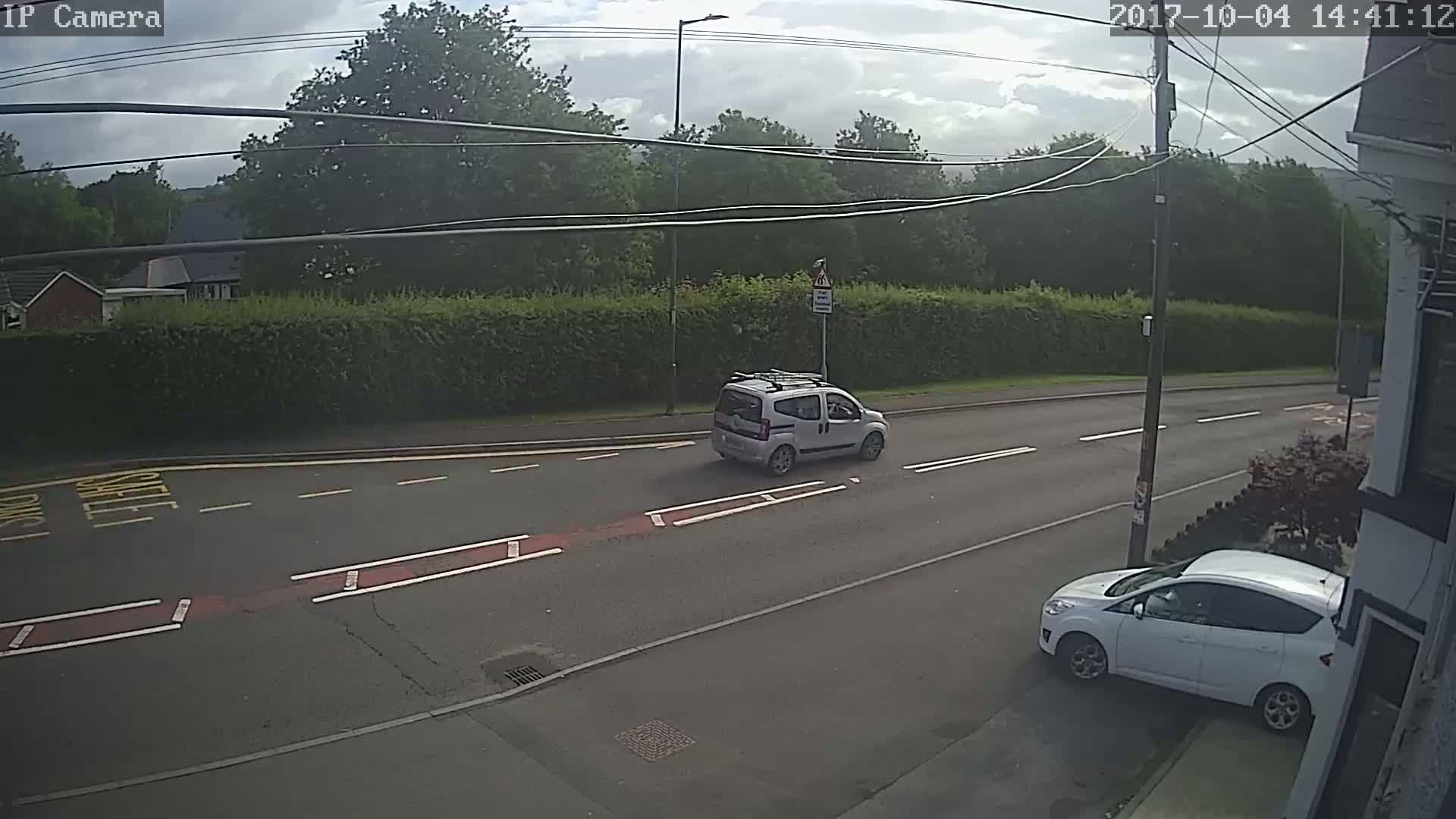 A light-grey car drives down a street lined with hedges under a partly cloudy sky.