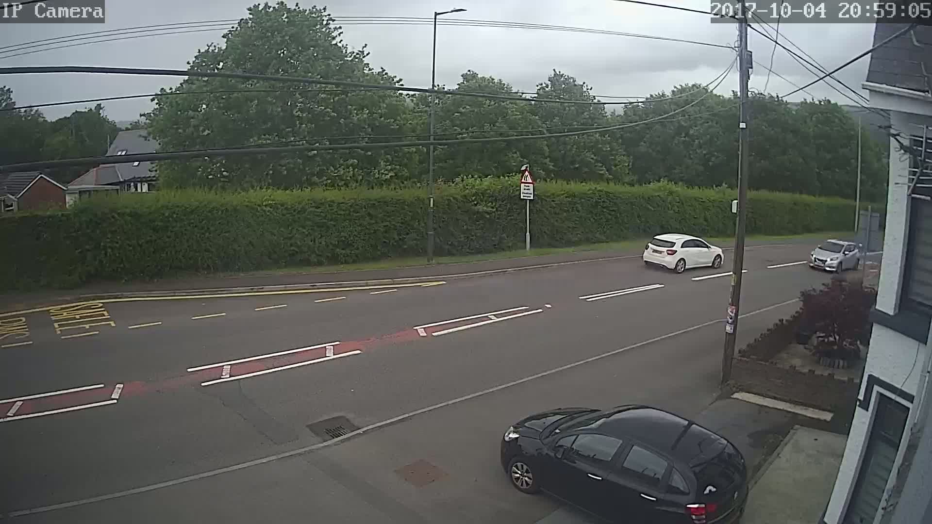 A street scene on an overcast day shows two cars driving on a road, and a third car parked near a house with a hedge in the background.