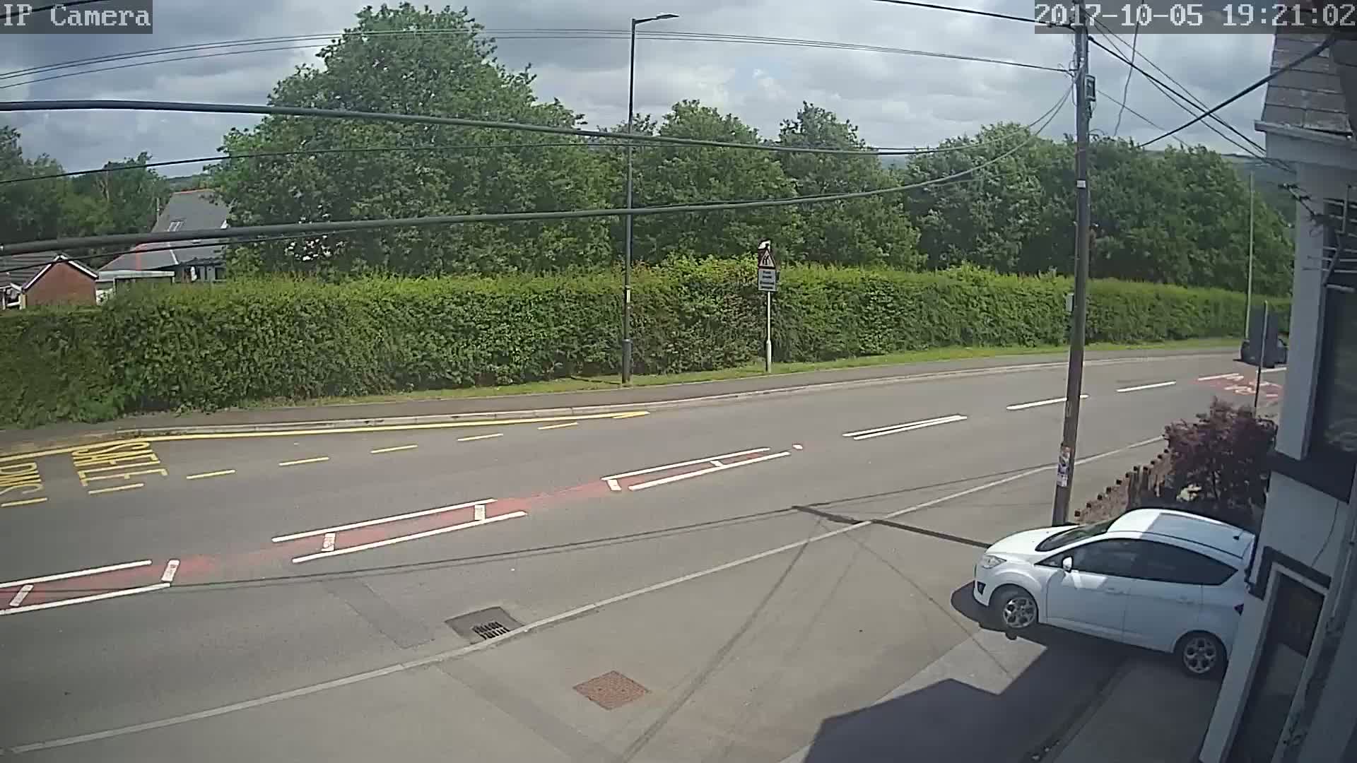 A partly cloudy day shows a white car parked on the side of a road next to a hedge, with other houses and trees visible in the background.