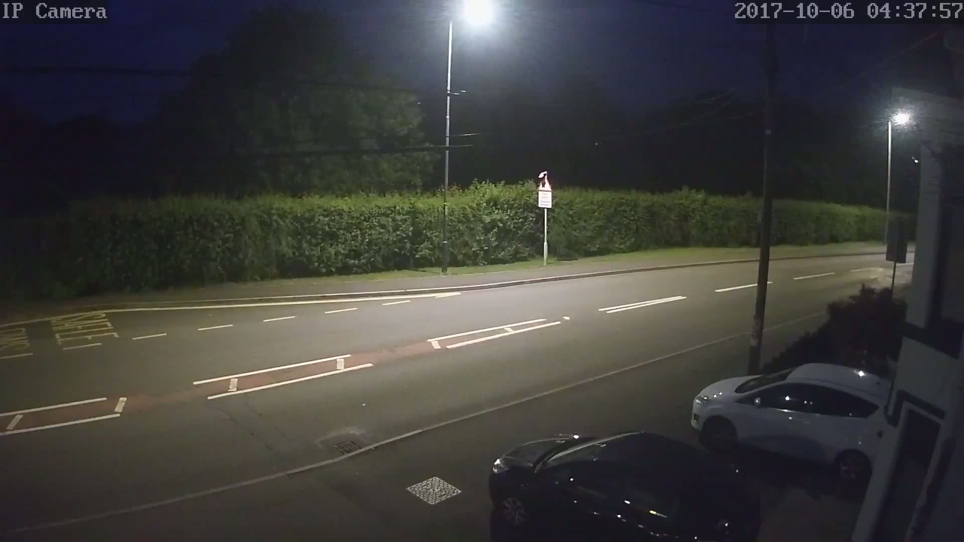 A nighttime street scene shows a road with parked cars and a hedge along the side, lit by streetlights.