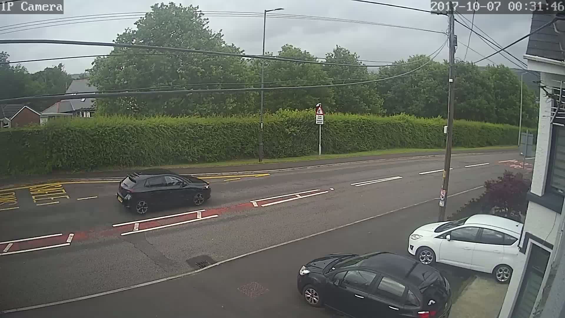 A dark-colored hatchback drives on a road lined with a tall hedge and trees under an overcast sky.