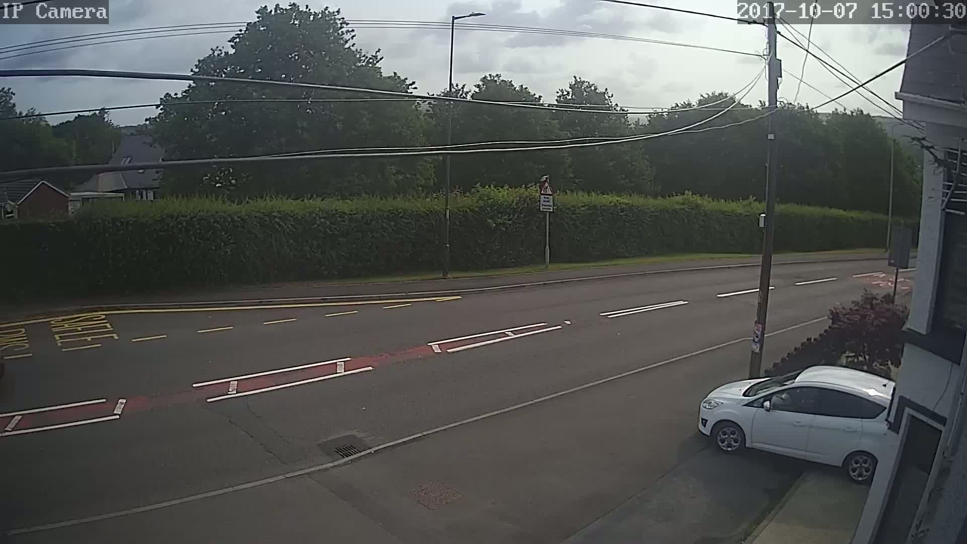 A white car is parked on the side of a road bordered by a tall hedge and trees under a partly cloudy sky.
