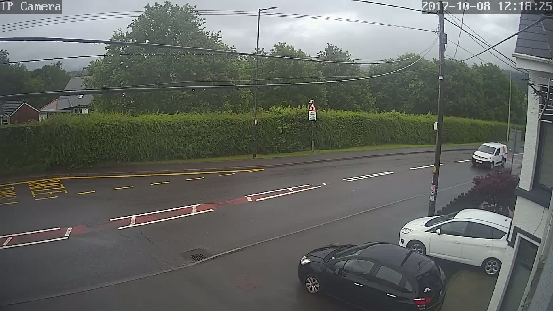 A wet, overcast street scene shows two cars parked and a van driving away, alongside a tall hedge and houses in the background.