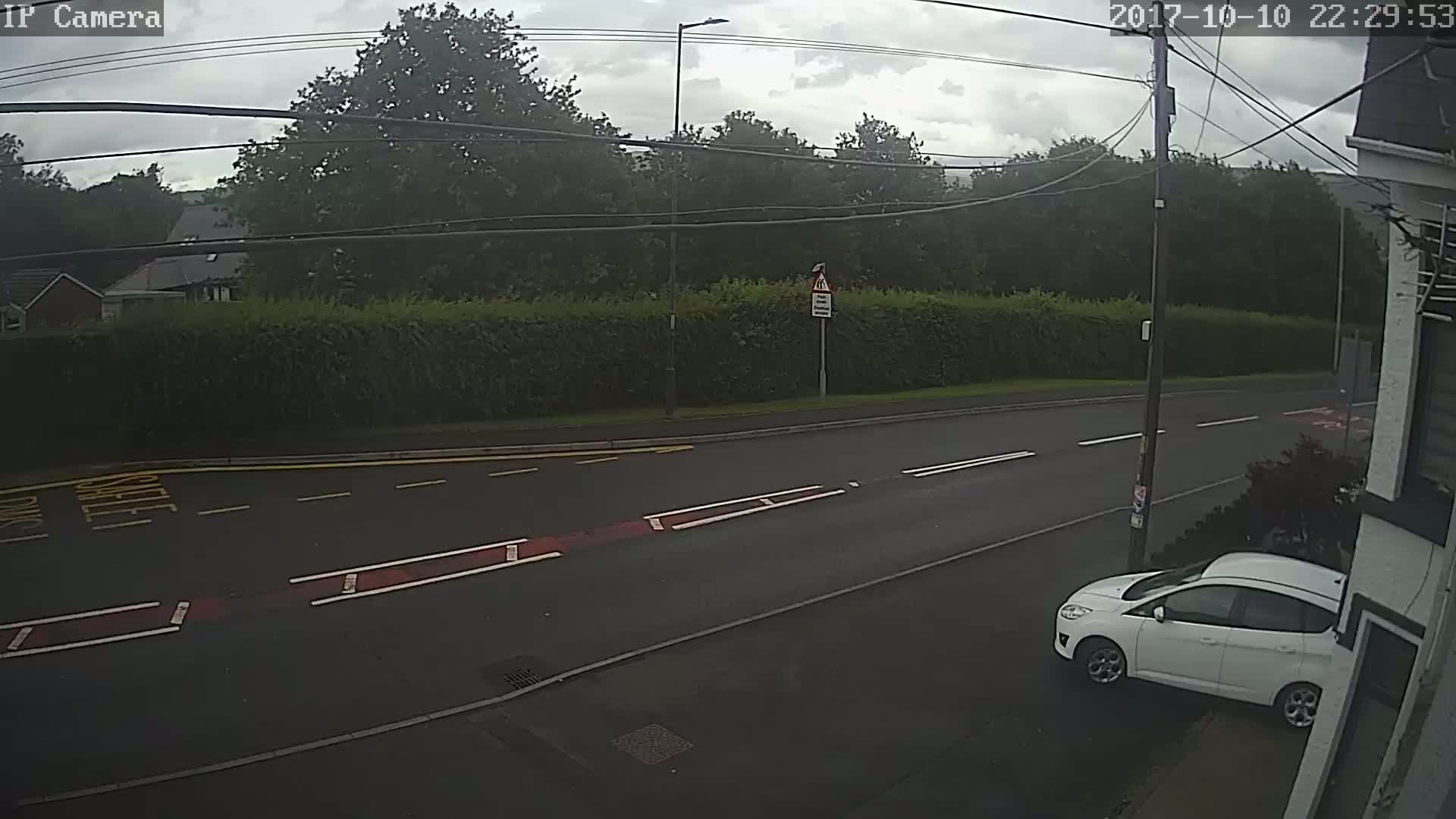 A white car is parked on the side of a road lined with green hedges under an overcast sky.