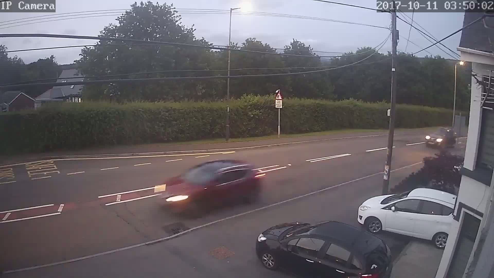 A dark red car drives past a white car parked on the side of a road under an overcast sky, with a black car partially visible in the lower right corner.