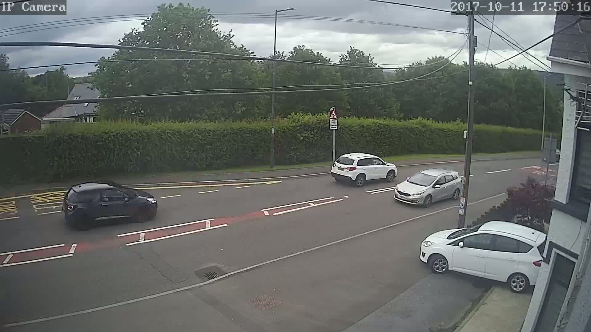 Three cars are driving on a road under a cloudy sky, near a hedge and houses.