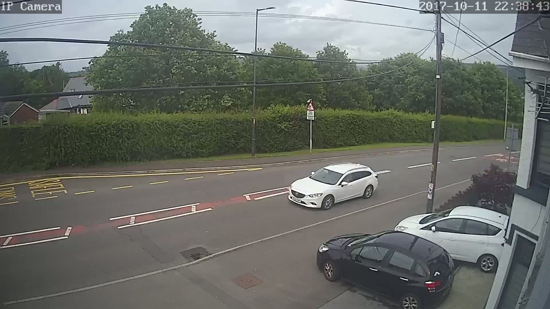 A white car drives down a road lined with hedges and trees under an overcast sky, with two other cars parked nearby.