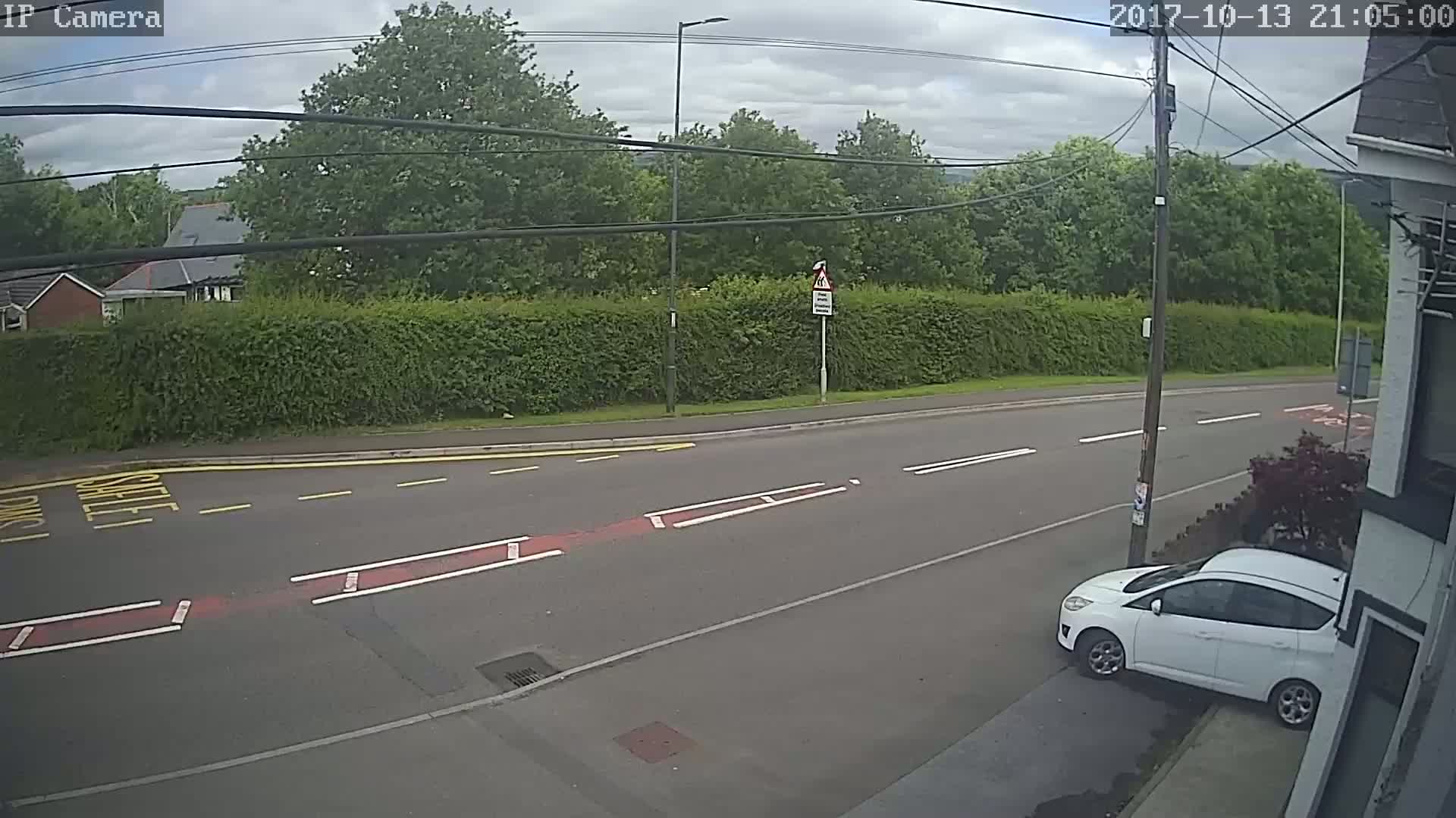 A mostly overcast day shows a white car parked on the side of a road, bordered by a tall hedge and trees, under a sky filled with clouds.