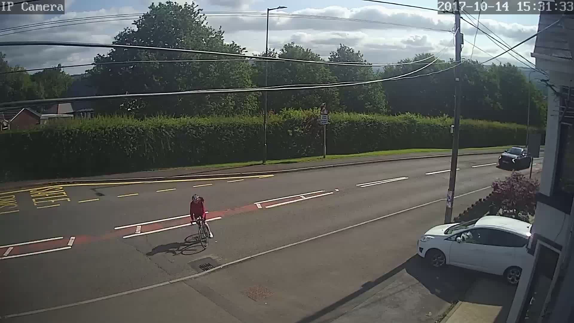 A cyclist in a red jacket rides a bicycle on a road under a partly cloudy sky, with a white car parked on the side of the road and another car further down the street.