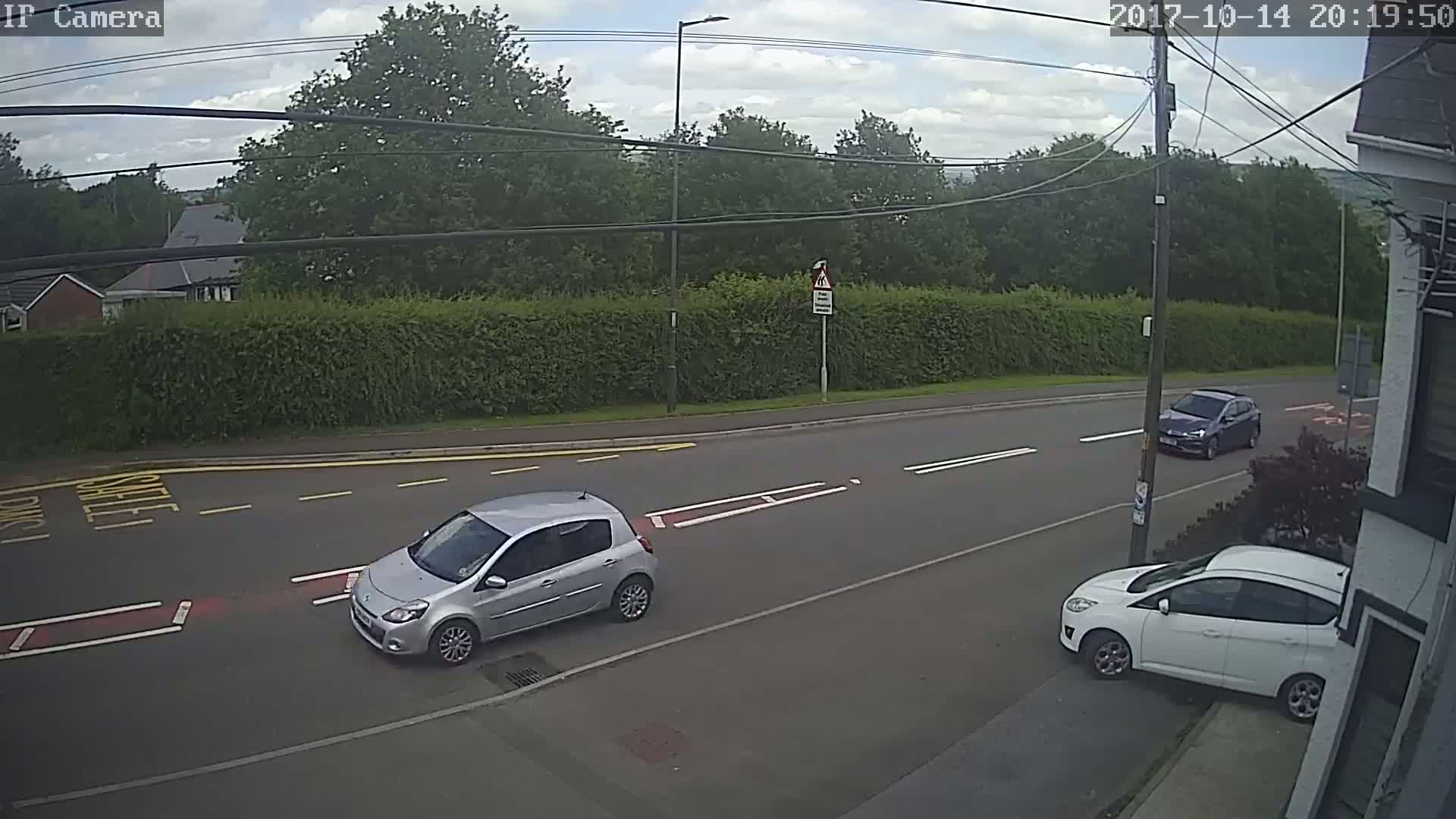 A partly cloudy day shows three cars on a road bordered by a tall hedge, with one car parked on the roadside.
