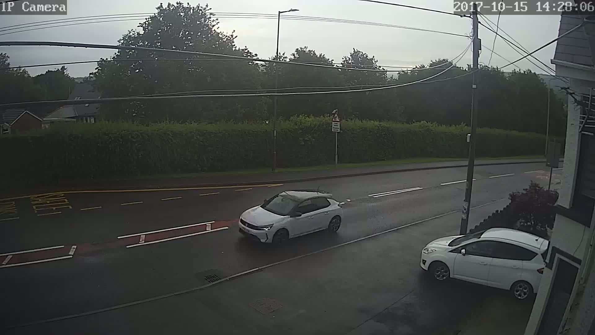 A wet road with two white cars, one driving and one parked, is seen under an overcast sky.