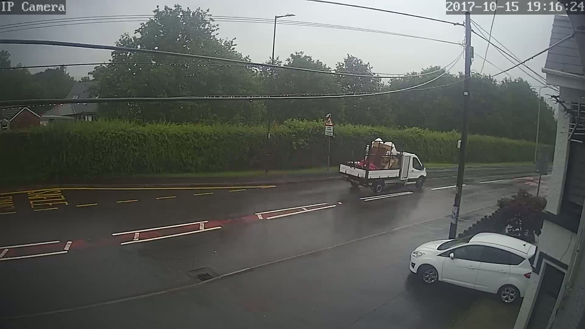 A white pickup truck carrying a load and a white car are parked on a rain-slicked road next to a hedgerow under a gray sky.