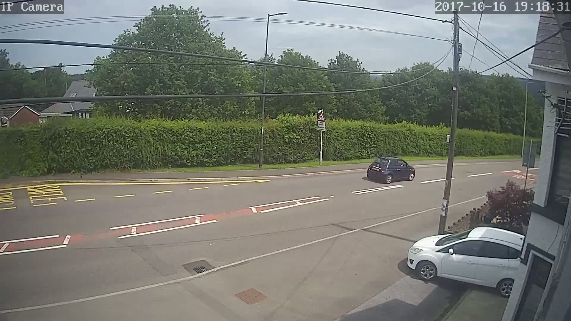 A mostly sunny day shows two cars, one dark-colored and one white, on a residential street with a tall hedge and trees in the background.