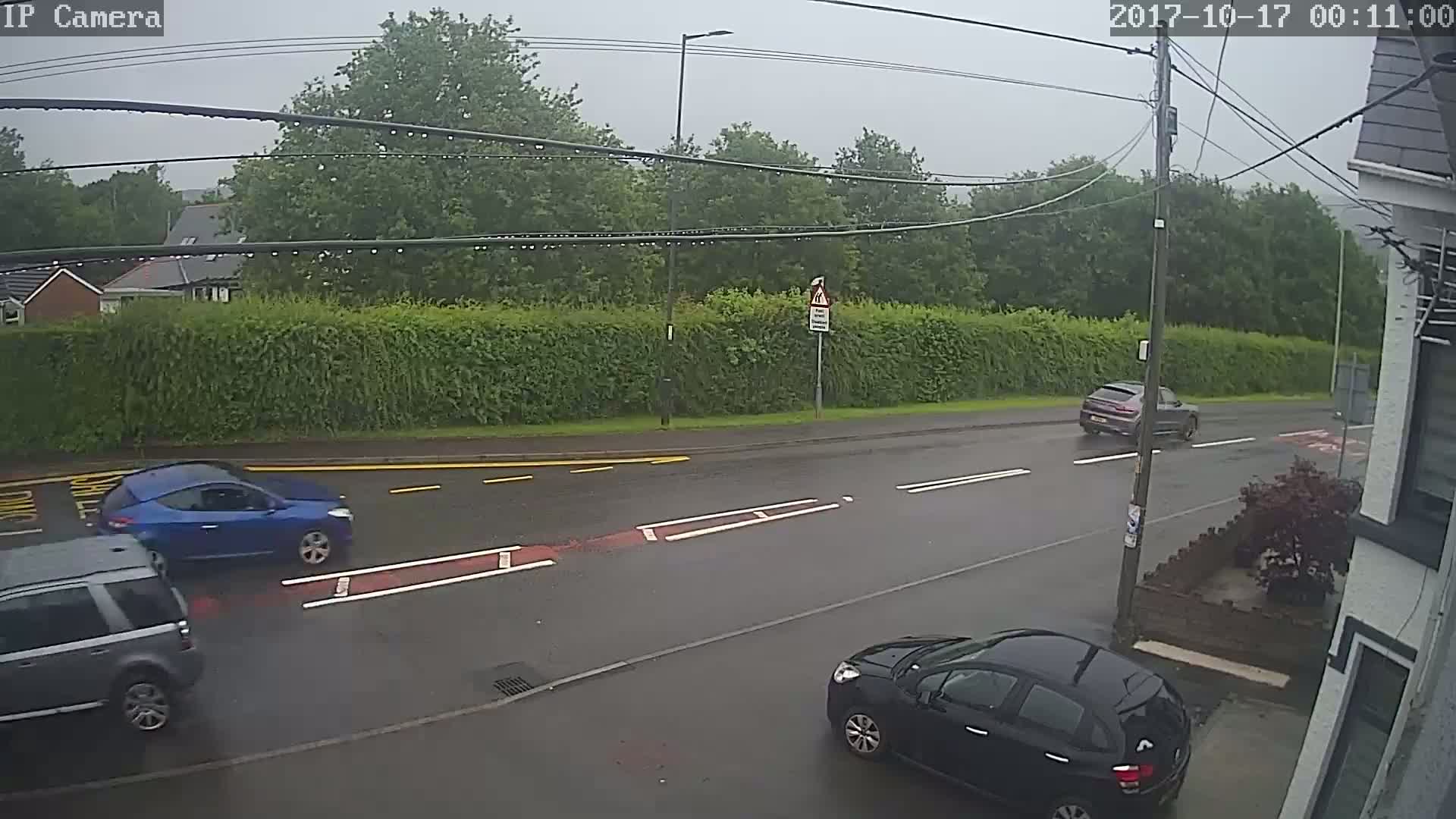 A rainy scene shows a street with several cars, including a blue car and a black car, and a hedge along the side of the road.