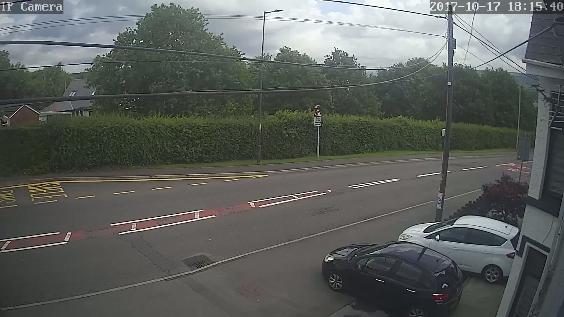 A mostly cloudy day shows two cars parked along the side of a road that runs past a tall hedge and line of trees.