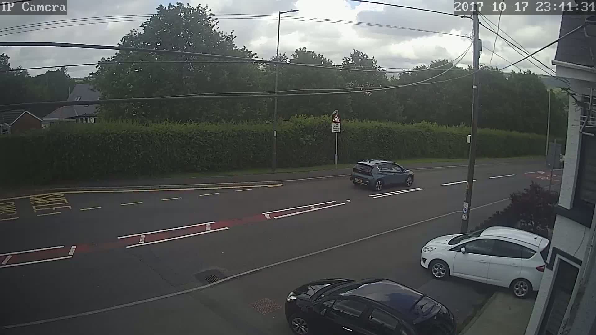 A partly cloudy day shows a road with three cars, one driving away and two parked, bordered by a tall hedge and trees.